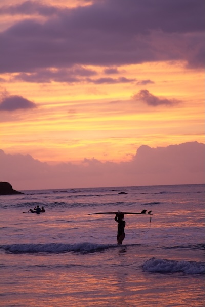 Femme tenant une planche de surf au-dessus de sa tête dans l’eau au coucher du soleil