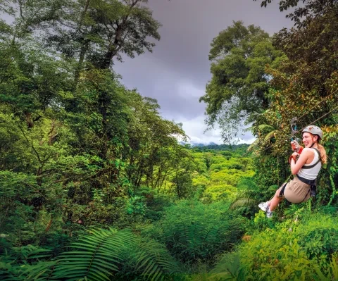 Un voyageur en tyrolienne à travers la canopée dense de la forêt tropicale costaricaine, suspendue au-dessus d’une végétation luxuriante avec des montagnes au loin.