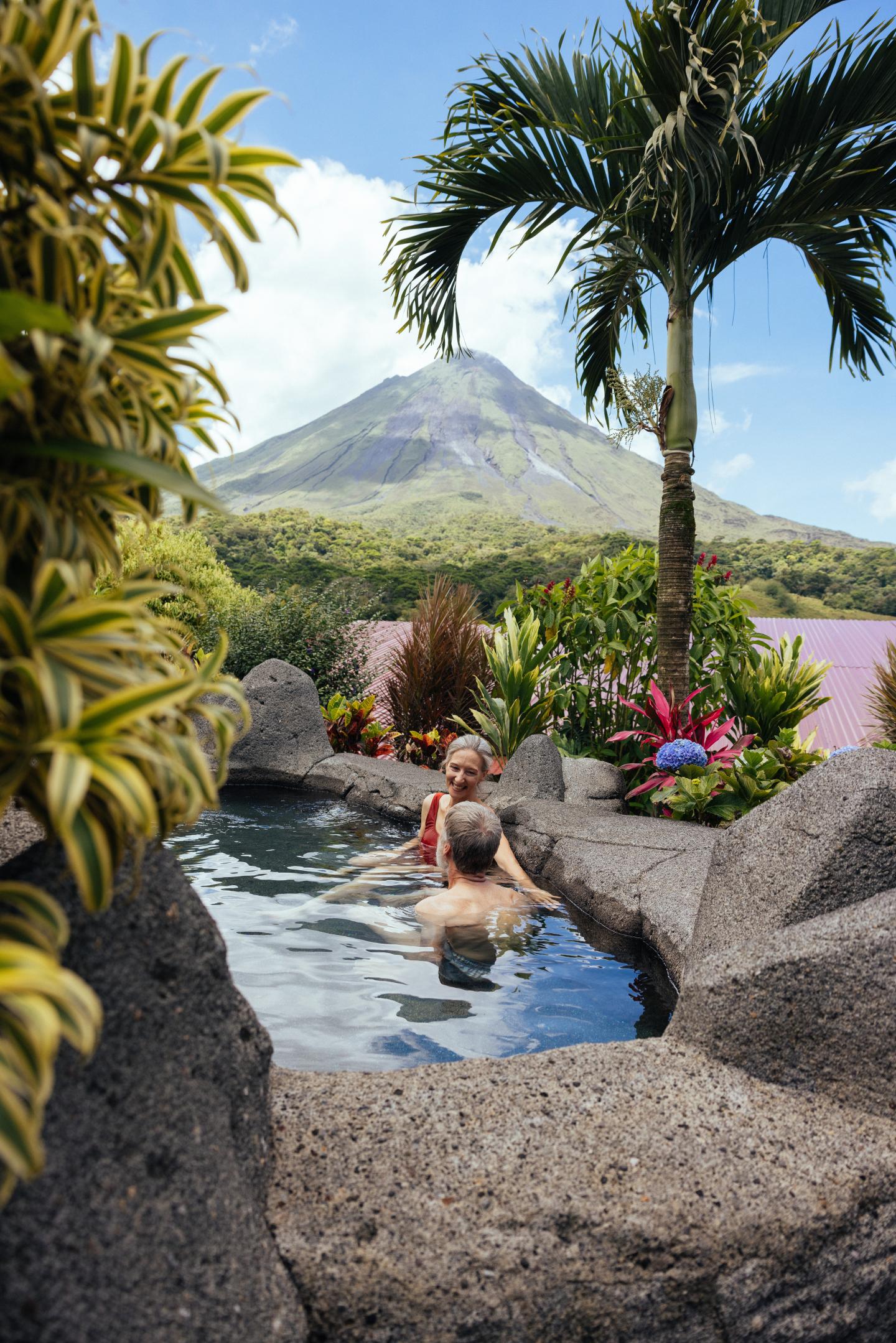 Deux personnes se détendent dans une piscine thermale en pierre entourée de plantes tropicales, avec le volcan Arenal en arrière-plan.
